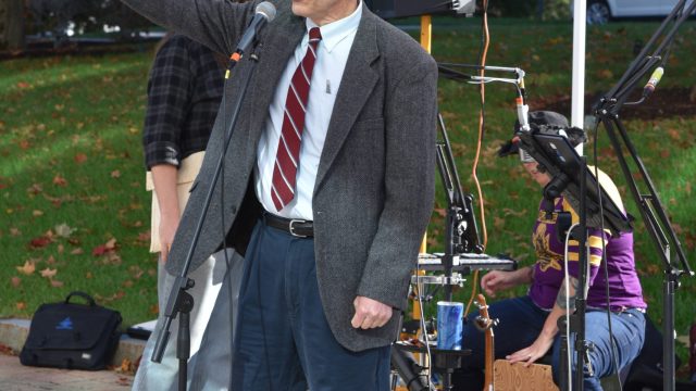 John Dieffenbacher-Krall, executive director of the Wabanaki Alliance, speaks to the crowd at the pre-march rally. Photo: Anne Crenshaw Man wearing a suit with red striped tie, glasses and outstretched hand speaks into a microphone
