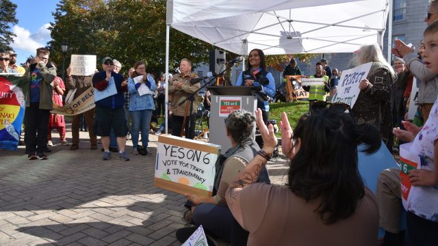 Maulian Dana, Penobscot Nation Ambassador and Wabanaki Alliance President, speaks to the crowd. Photo: Anne Henshaw Woman stands at podium under white tent before a crowd holding signs that read Yes on 6 Vote for Truth and Transparency
