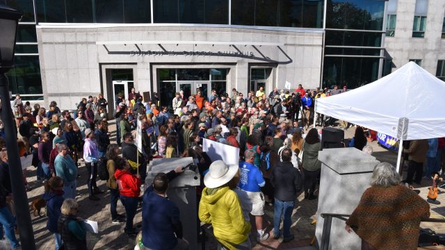 More than 200 people attended the rally and then joined a march around the State House. Photo: Anne Henshaw Crowd of people in front of grey building with white tent in foreground